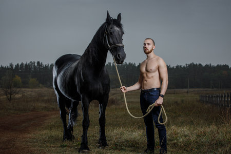 Young bearded man with naked torso standing with horse at the countrysideの写真素材