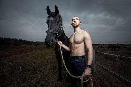 Young strong man with naked torso standing with horse at the countrysideの写真素材