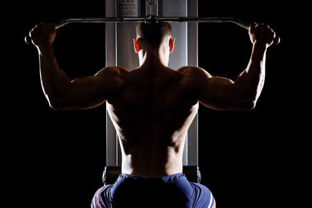 Silhouette of young handsome topless bodybuilder works out pulling down on cable machine in gym.