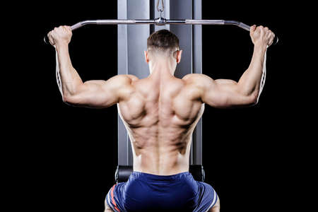 Back view of young handsome topless bodybuilder works out pulling down on cable machine in gym.の写真素材