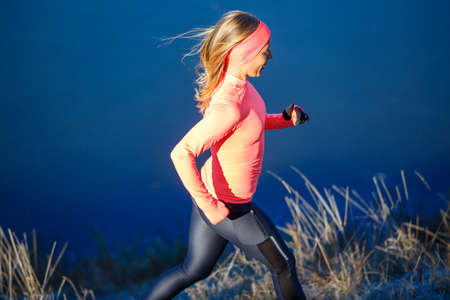 Young sporty woman in orange longsleeve running along blue frosty lake in the morningの写真素材