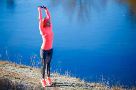 Young slim fitness woman stretching near lake before jogging in the frosty morning.