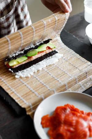 Young woman wrapping up sushi roll ingredients on bamboo matの写真素材