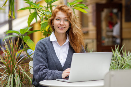 Young red-haired smiling woman using laptop in cafe outdoorの写真素材