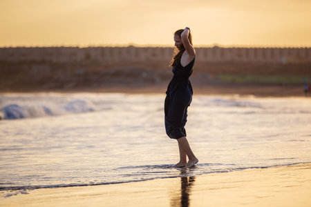 Young beautiful woman in long dress walking at the beach at sunsetの写真素材
