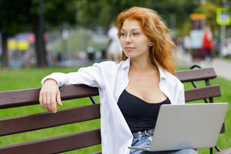 Relaxing young redhead woman sitting in the park and using her laptopの写真素材