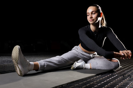 Young fitness girl stretching her legs on the mat in the gym isolated on blackの写真素材