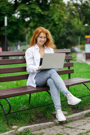 Young confident attractive student girl using her laptop sitting on the bench in parkの写真素材