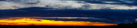 Panoramic sunset sky with heavy dark blue clouds and orange line of sunの写真素材