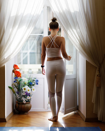 Young slim woman in white sportswear standing near open balcony in the morning