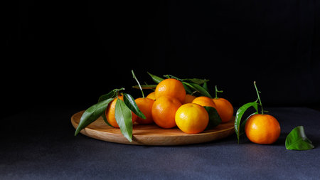 Juicy tangerines with vibrant leaves arranged on a wooden plate against a dark background.の写真素材