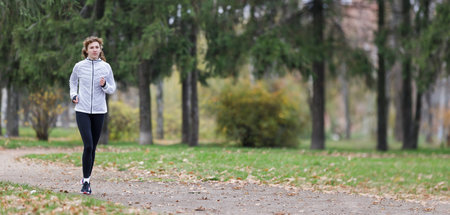 A woman is jogging on a gravel path surrounded by trees in a park, with colorful autumn leaves scattered on the ground. The fresh air and peaceful environment enhance her workout routine.の写真素材