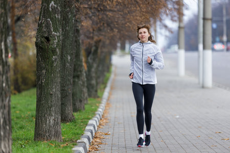 A woman dressed in a light jacket and leggings jogs along a pathway bordered by trees. The early morning atmosphere features a slight fog.の写真素材