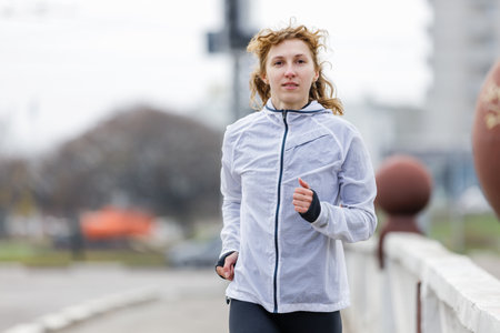 A young woman jogs energetically along an urban pathway wearing a light jacket. Her focused expression shows determination as she enjoys an outdoor exercise session on a cloudy day.の写真素材