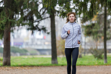 A young woman runs energetically along a gravel path in a vibrant park, surrounded by tall trees....
