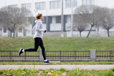 A young woman runs at a steady pace on a park path surrounded by foggy weather. The serene atmosphere and her focused expression highlight her dedication to fitness in nature.の写真素材
