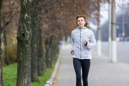 A woman dressed in a light jacket and leggings jogs along a pathway bordered by trees. The early morning atmosphere features a slight fog.の写真素材