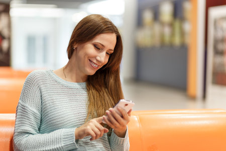 Young woman sitting on an orange bench using a smartphone in a modern, well-lit indoor space during daytime.の写真素材