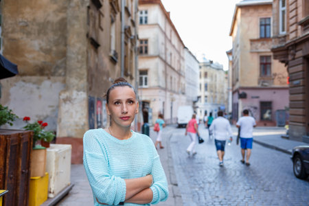 A young woman in a light sweater stands with her arms crossed in a charming cobblestone street, surrounded by historic buildings and passing pedestrians enjoying the day.の写真素材