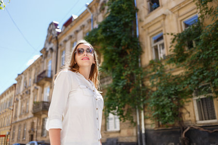 Smiling young woman enjoying a sunny day in a historic urban setting with classic architecture and lush greenery in the backgroundの写真素材
