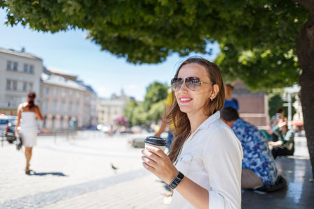 A young woman enjoys her coffee on a sunny day in a vibrant city square, surrounded by greenery and lively street activity.の写真素材