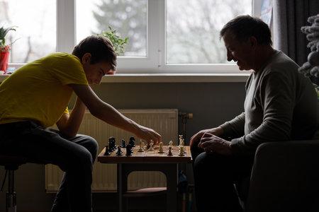 Young teenage boy and older man enjoying a chess game indoors during daylight hoursの写真素材