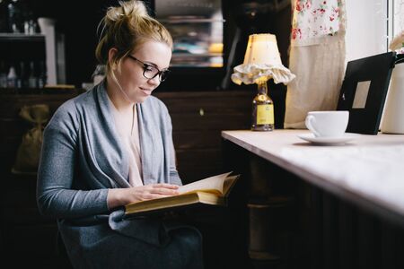 beautiful young woman reading interesting book while drinking tea at home, in cafe