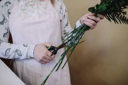 Woman florist in apron cuts off stems of flowers indoor. Female florist preparing bouquet in flower shop. Close upの写真素材