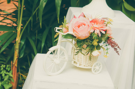 Mixed artificial flowers in small white bicycle vase and basket on white table in garden. Vintage style.の写真素材