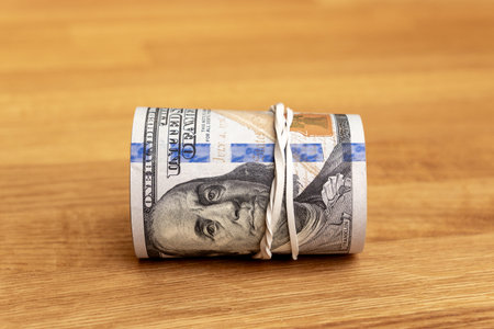 Close-up of a roll of American one hundred dollar bills tied with a white rubber band, isolated on a textured wooden table surface. Concept of savings, wealth, cash storage, or financial success.