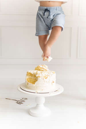 Baby boy feet in blue shorts on the first birthday cake on a white wooden background. Celebration and funの写真素材