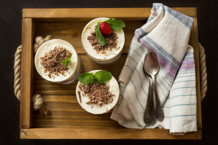 White dessert with a layer of strawberries in a transparent glass with sprinkled with grated chocolate and a mint leaf on a wooden tray with a natural kitchen napkin and teaspoons. Flatlayの写真素材