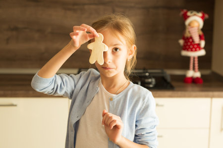 A girl in a blue sweater and a white T-shirt holds a Christmas man biscuit in her hands at face level against the background of the kitchen and a New Year's doll. Homemade baking conceptの写真素材