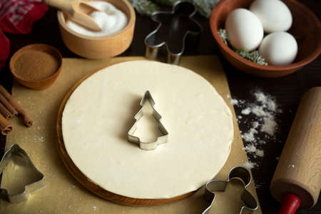 Culinary Christmas background with baking ingredients - round sheet of dough on the podium, cookie cutters, rolling pin, sugar, eggs, dried orange, two kinds of cinnamon, parchment, apron. Flat layの写真素材