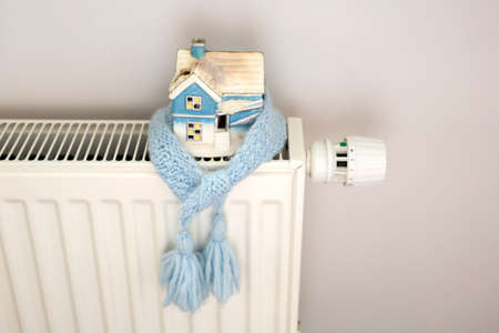 White and blue house model wrapped in blue knitted scarf with tassels on a radiator indoors. Winter heating season concept. Front view. Space for textの写真素材