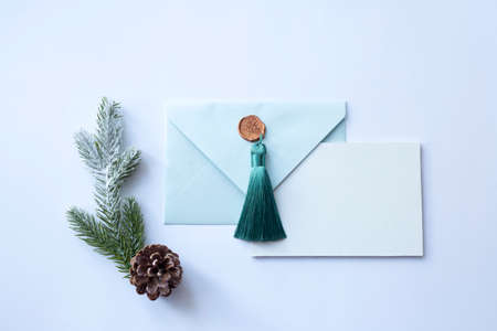 Turquoise envelope with a beautiful gold tassel and a white sheet of paper on a white background with a sprig of a Christmas tree and a pine cone. Flat lay. Place for textの写真素材