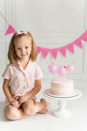 A cute little girl in pink pajamas and a bow on her head next to her birthday cake decorated with pink balloons. On a white background with pink holiday flags.の写真素材