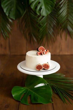 White cake with fig decor on a wooden white stand on a brown background. Decorated with palm leaves and monsteraの写真素材