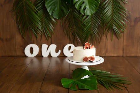 White cake with fig decor on a wooden white stand on a brown background. Decorated with palm leaves and monstera. Letters oneの写真素材
