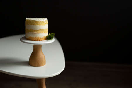 A tiny naked cake on a wooden stand with a brown base on a white table against a black background....