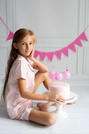 A beautiful girl with long hair in pink pajamas with a birthday cake decorated with balloons, on a stand on a white background with pink flags.の写真素材