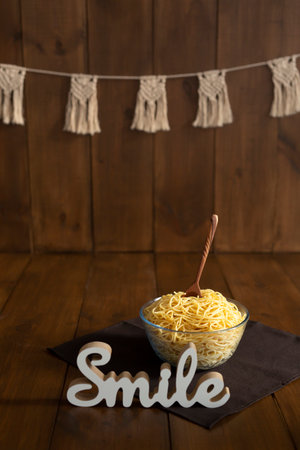 A transparent bowl of pasta with a wooden spoon inside on a wooden braun background with a white figurine Smile next to it. Spaghetti smash conceptの写真素材