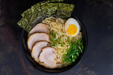 Top view of a bowl of ramen noodles with meat, egg, and greens on dark background.の素材