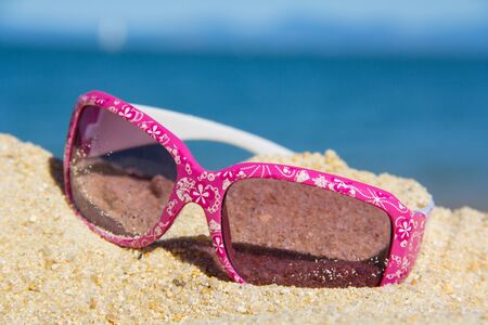 pink kids suglasses with a white floral design on the sand near the seaの写真素材