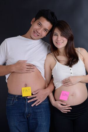 Belly of asian pregnant wife and husband with funny inscription "FAT" and "SON"の写真素材