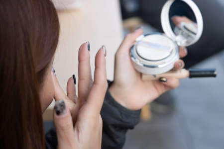 lady doing makeup her left hand holds a powder foundation and mascara while her right hand touches her face.の写真素材