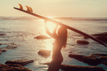 Silhouette surfer girl in bikini on tropical beach holding surfboard at sunset