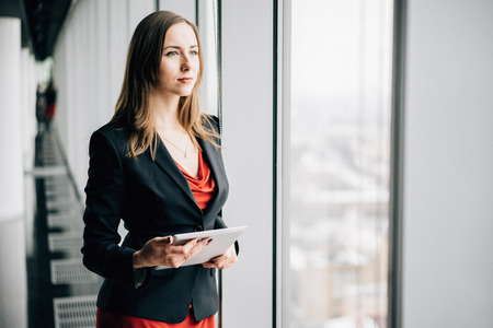Young business woman in a red dress and jacket, standing near the window, winter city landscape outside the window on the backgroundの写真素材