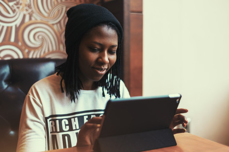 Smiling young happy black african american girl sitting on a leather sofa in a cafe at daytime, wearing a hat and long sleeve shirt, with a tablet, vintage color filterの写真素材