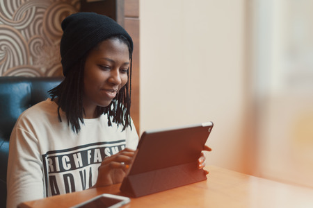 Smiling young happy black african american girl sitting on a leather sofa in a cafe at daytime, wearing a hat and long sleeve shirt, with a tablet and smartphone, nail art, vintage color filterの写真素材